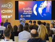 Audience watches a speaker beneath a screen promoting audio radio content Audience watches a speaker beneath a screen promoting audio radio content